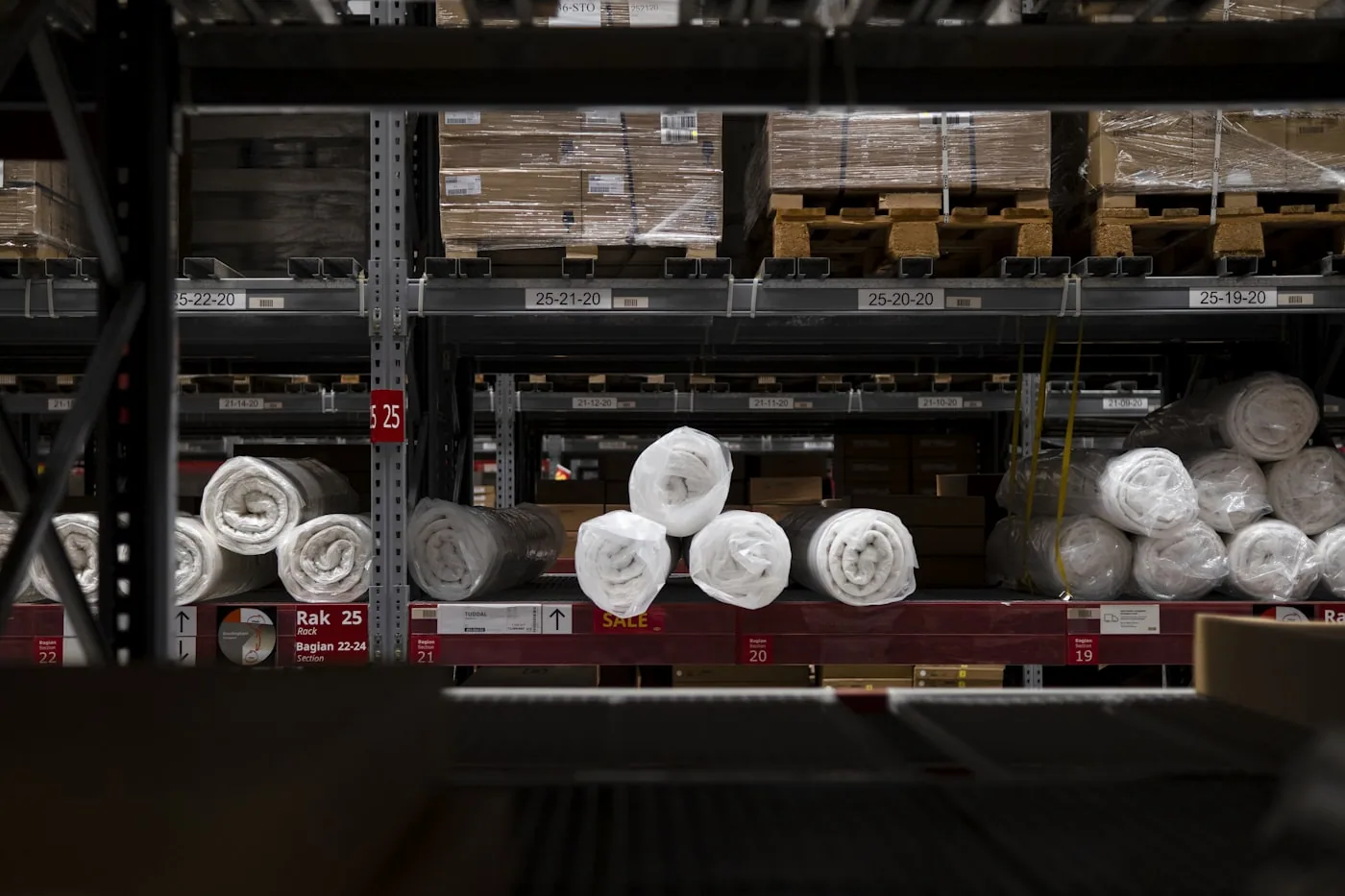 Rouleaux de tissus étiquetés par coloris et laize empilés dans un entrepôt textile.