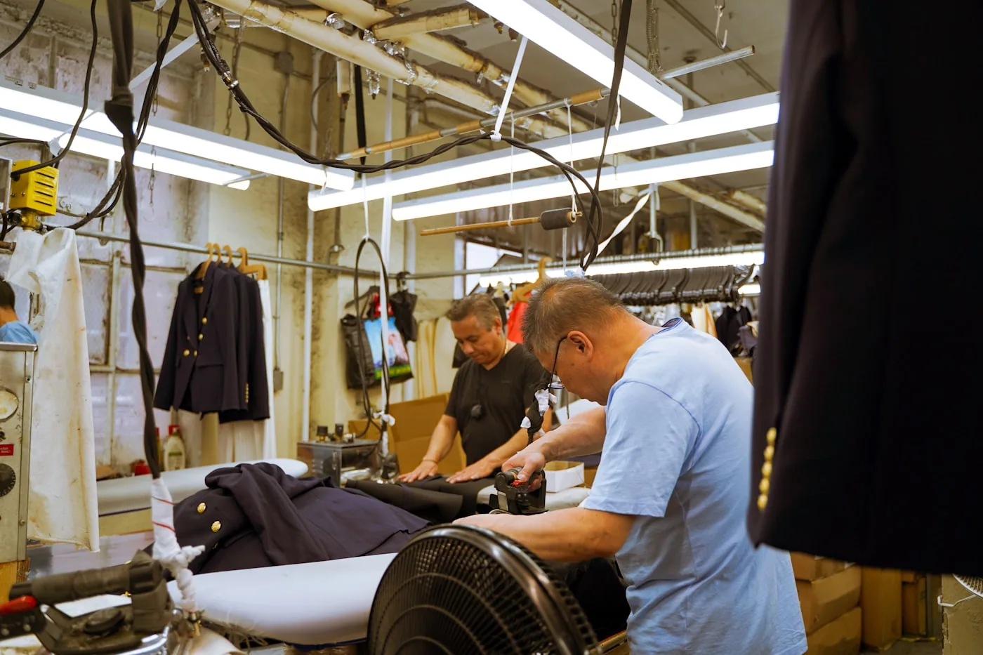 Responsable de marque et chef d’atelier discutant au milieu d’une ligne de confection, avec postes de couture et rouleaux de tissu en arrière-plan.
